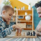Smart child playing chess with dad at home lying on floor together having fun with intellectual sports game. Happy family and leisure activity concept.