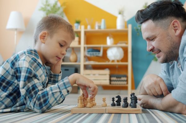Smart child playing chess with dad at home lying on floor together having fun with intellectual sports game. Happy family and leisure activity concept.