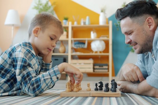 Smart child playing chess with dad at home lying on floor together having fun with intellectual sports game. Happy family and leisure activity concept.
