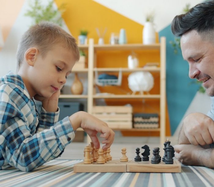 Smart child playing chess with dad at home lying on floor together having fun with intellectual sports game. Happy family and leisure activity concept.
