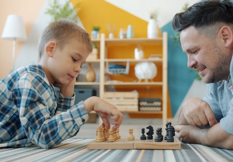 Smart child playing chess with dad at home lying on floor together having fun with intellectual sports game. Happy family and leisure activity concept.